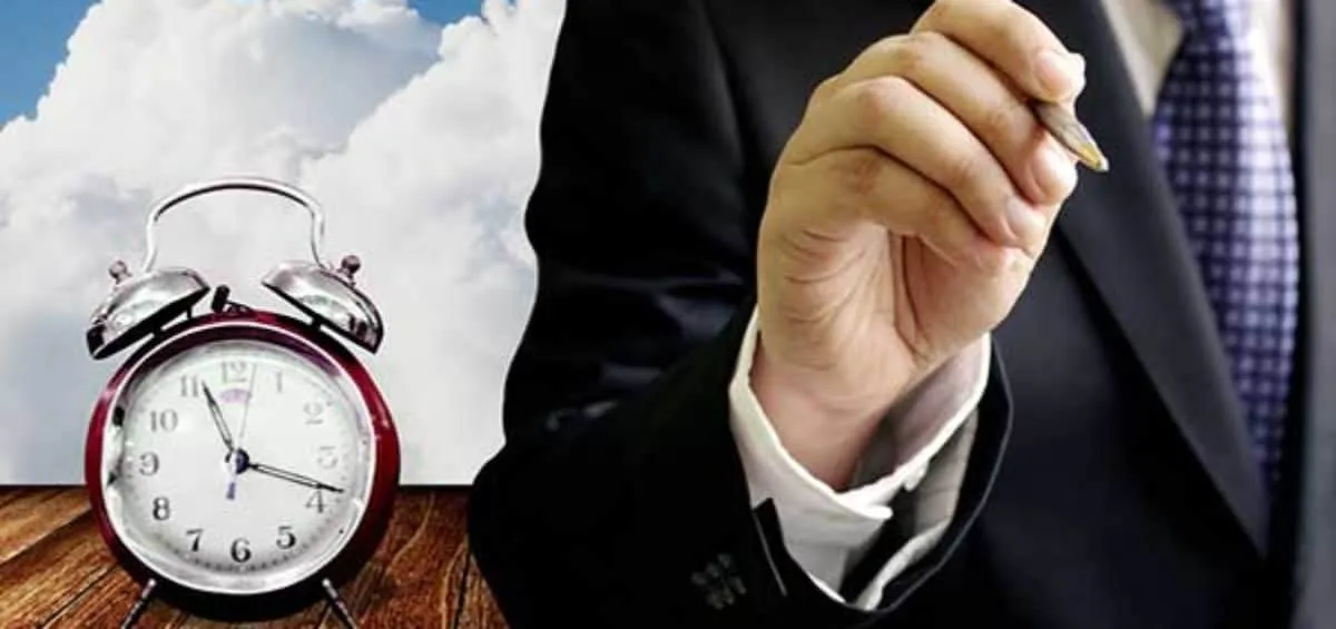 Una sveglia da tavolo accanto alla mano di un uomo in giacca che regge una penna
